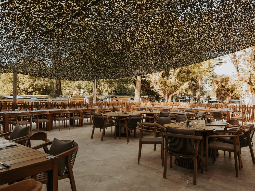 La grande terrasse de L'Espagnette sous la voile, tables et fauteuils en bois à l'ombre des arbres